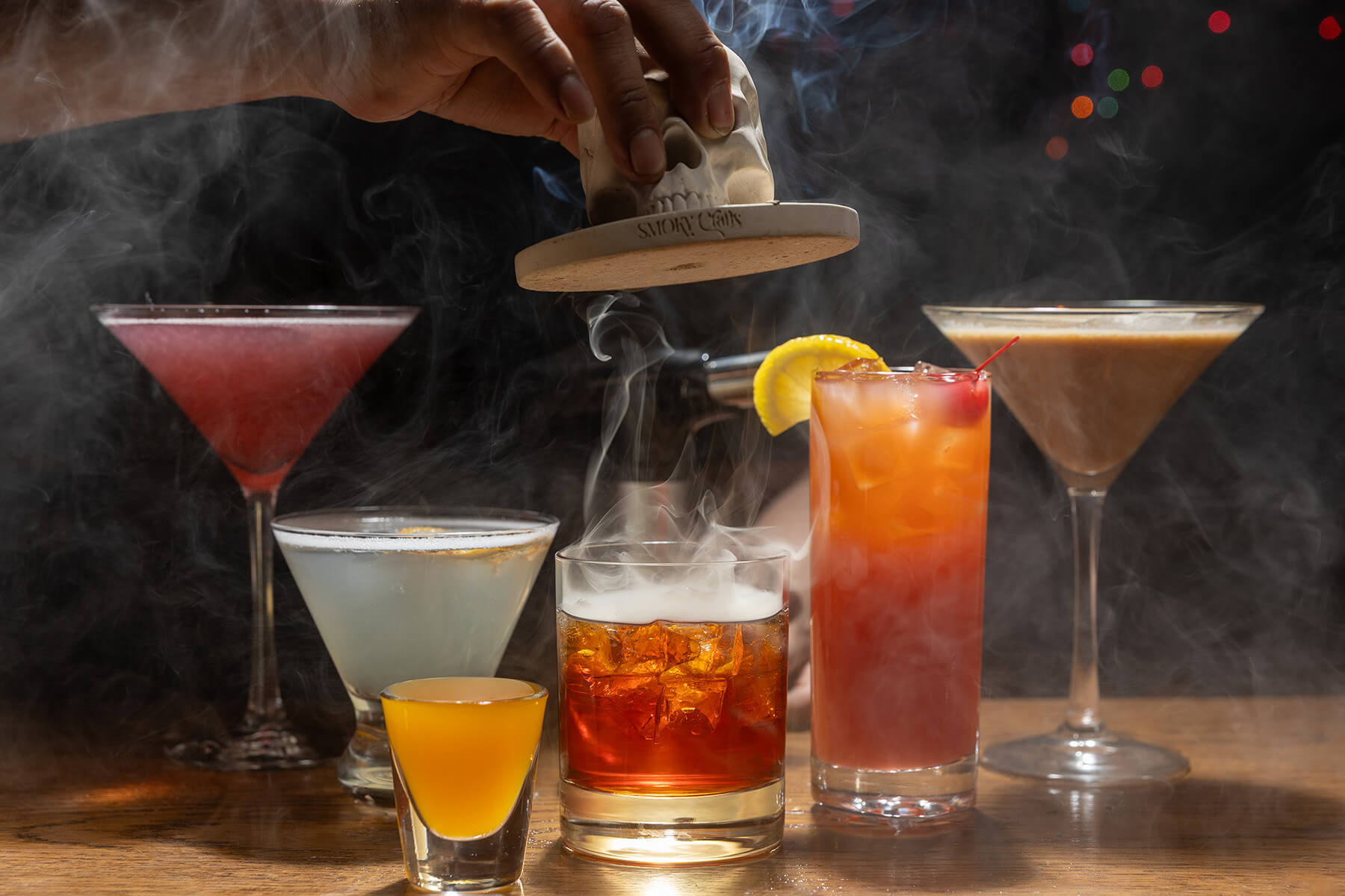 Various colorful cocktails on a wooden table surrounded by smoke, with a hand lifting a skull-shaped lid releasing smoke.